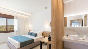 A modern bedroom with a large bed, a bedside table, a nightstand, a window with curtains, a bathroom with a sink and mirror, and a view of a desert landscape. - Villa Santa Irini 3 - With Heated Pool (Agia Eirini)