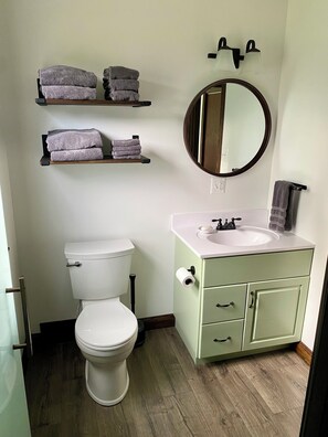 A bathroom with a toilet, a sink, a mirror, and shelves with towels. - Cozy Sandhill Cottage, 15mi from Crystal Mountain, dog friendly, AC
(Elberta)