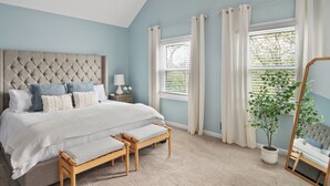 A bedroom with a tufted headboard, a large bed with pillows, a bedside table with a lamp, a window with blinds, a potted plant, and a mirror.
