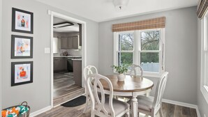 A dining area with a wooden table and white chairs, a window with a view of trees, and a kitchen in the background.