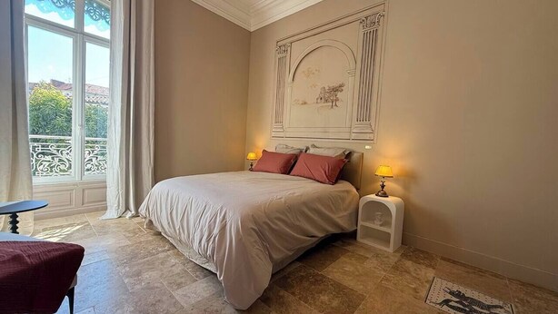 A bedroom with a bed, two bedside lamps, a small table, and a balcony view. - To complete (Nîmes)