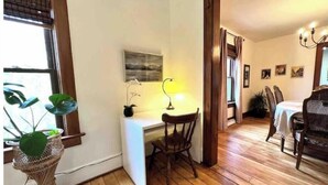 Ein Zimmer mit Holzboden, einem Schreibtisch mit Lampe, einem Stuhl, einer Topfpflanze und einem Essbereich mit Stühlen und Tisch.