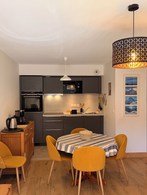 Een compacte keuken met moderne apparaten, een eettafel met stoelen en een bijzondere lichtarmatuur met een uniek patroon.