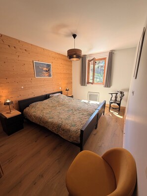 A bedroom with a wooden wall, a bed, a nightstand with a lamp, a chair, and a window with curtains.