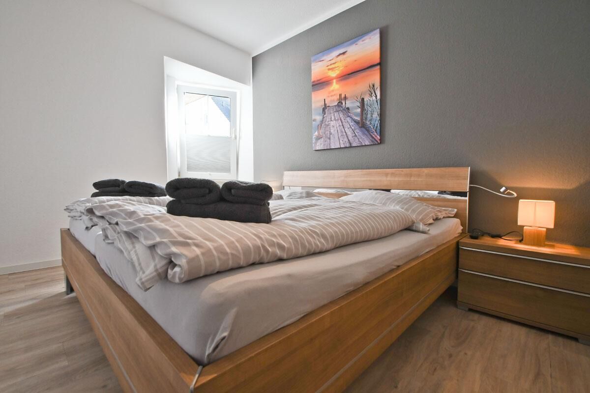 Une chambre moderne avec un lit en bois, une table de chevet avec une lampe et un tableau représentant un coucher de soleil accroché au mur.