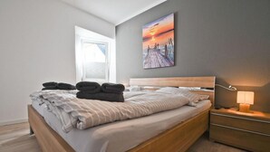 A modern bedroom with a wooden bed, a nightstand with a lamp, and a painting of a sunset on the wall. - Vacation apartment Wellenreiter (Norderney)