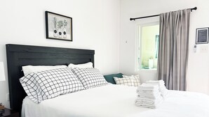 A neatly made bed with white linens and checkered pillows, a framed botanical illustration on the wall, and a window with curtains.