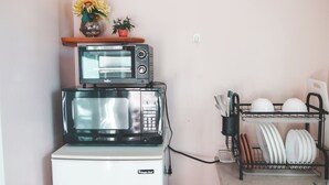 A compact kitchen with a microwave oven stacked on top of a smaller appliance, a dish rack with plates, and a vase with flowers on a shelf.