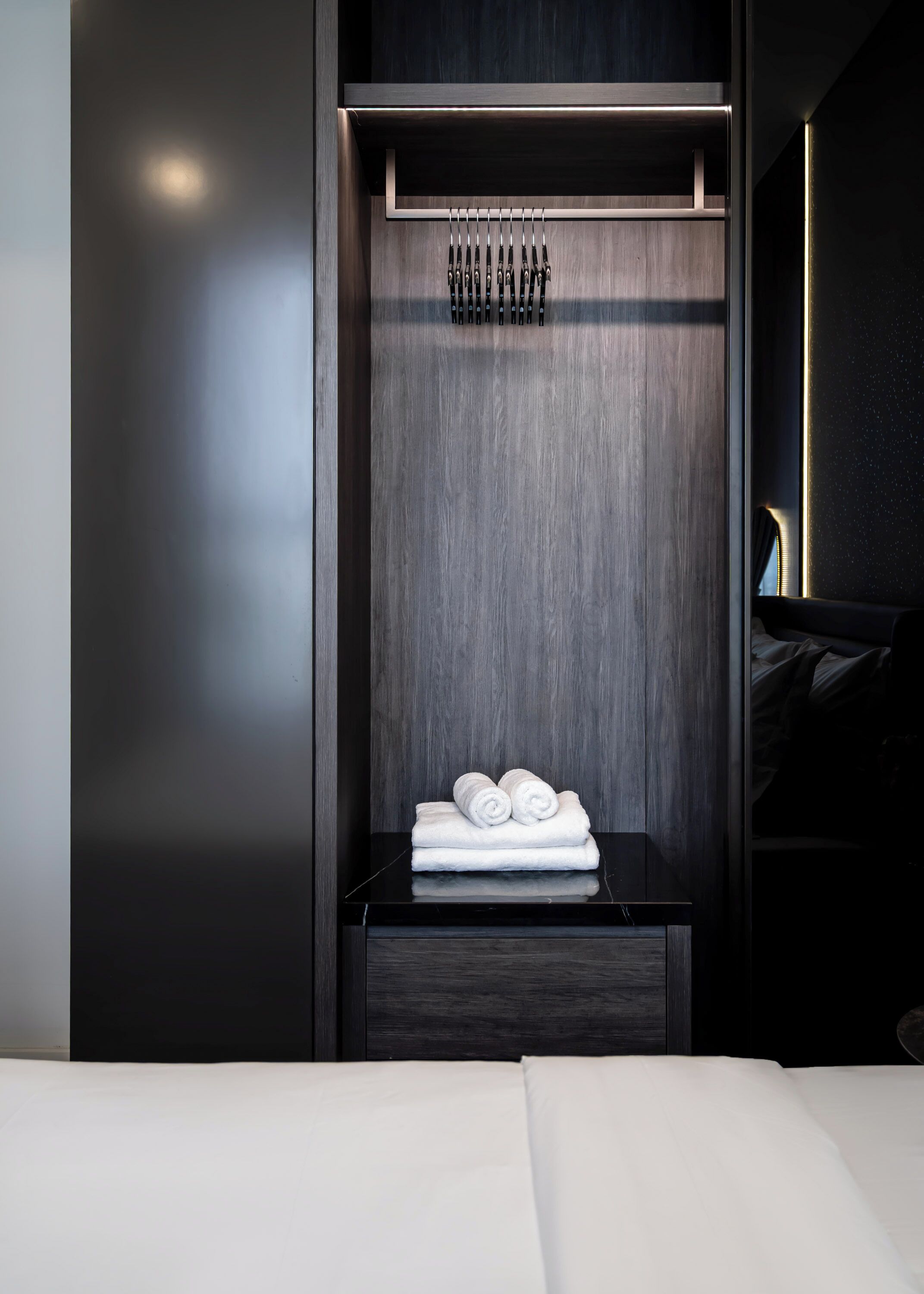 Ein modernes Hotelzimmer mit einem dunklen Holzschrank, einem ordentlich bezogenen Bett und einem kleinen Nachttisch mit gerollten Handtüchern.