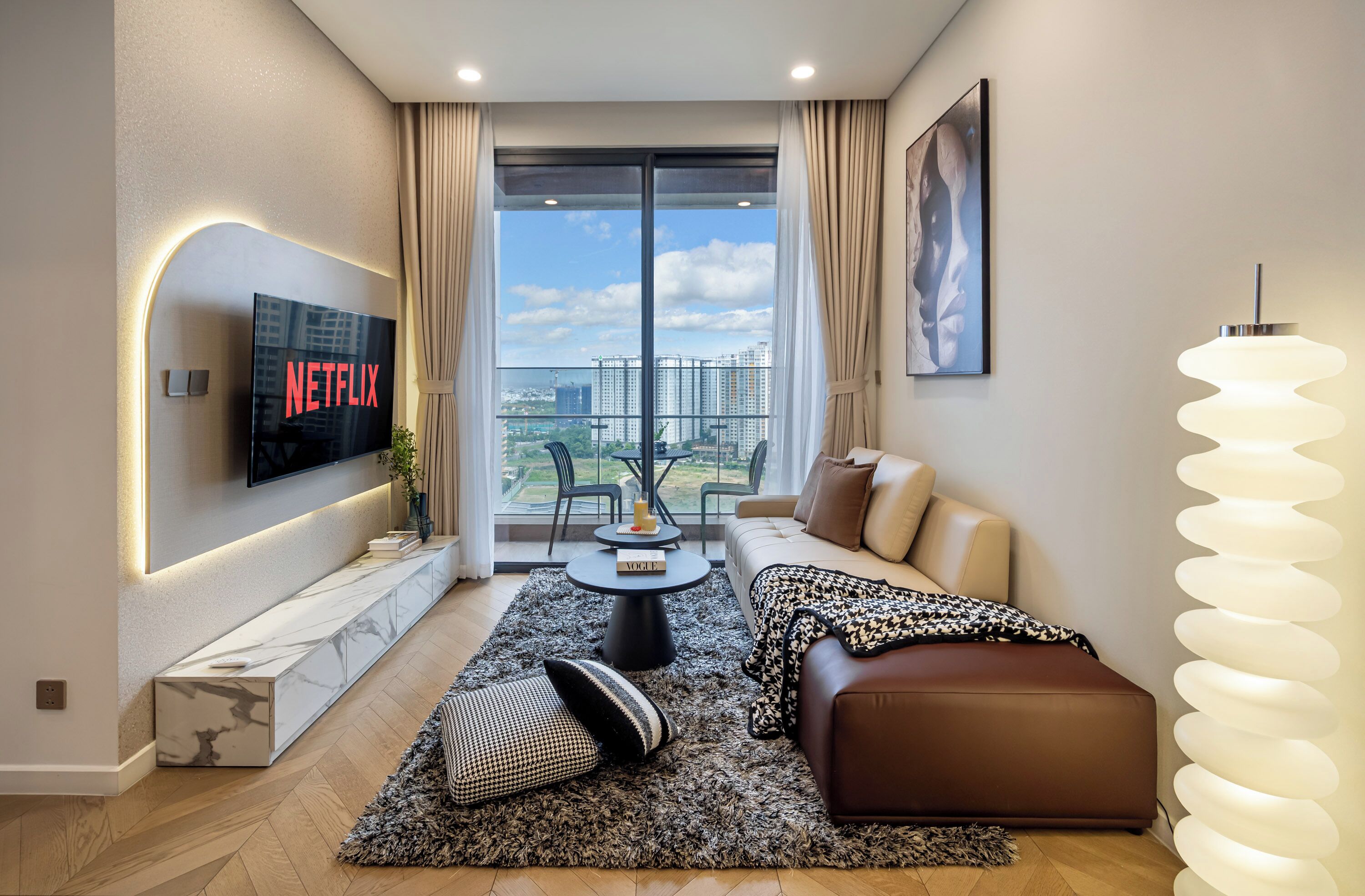 En moderne stue med et stort TV, der viser 'NETFLIX', en sofa, et sofabord og udsigt over en by gennem store vinduer.