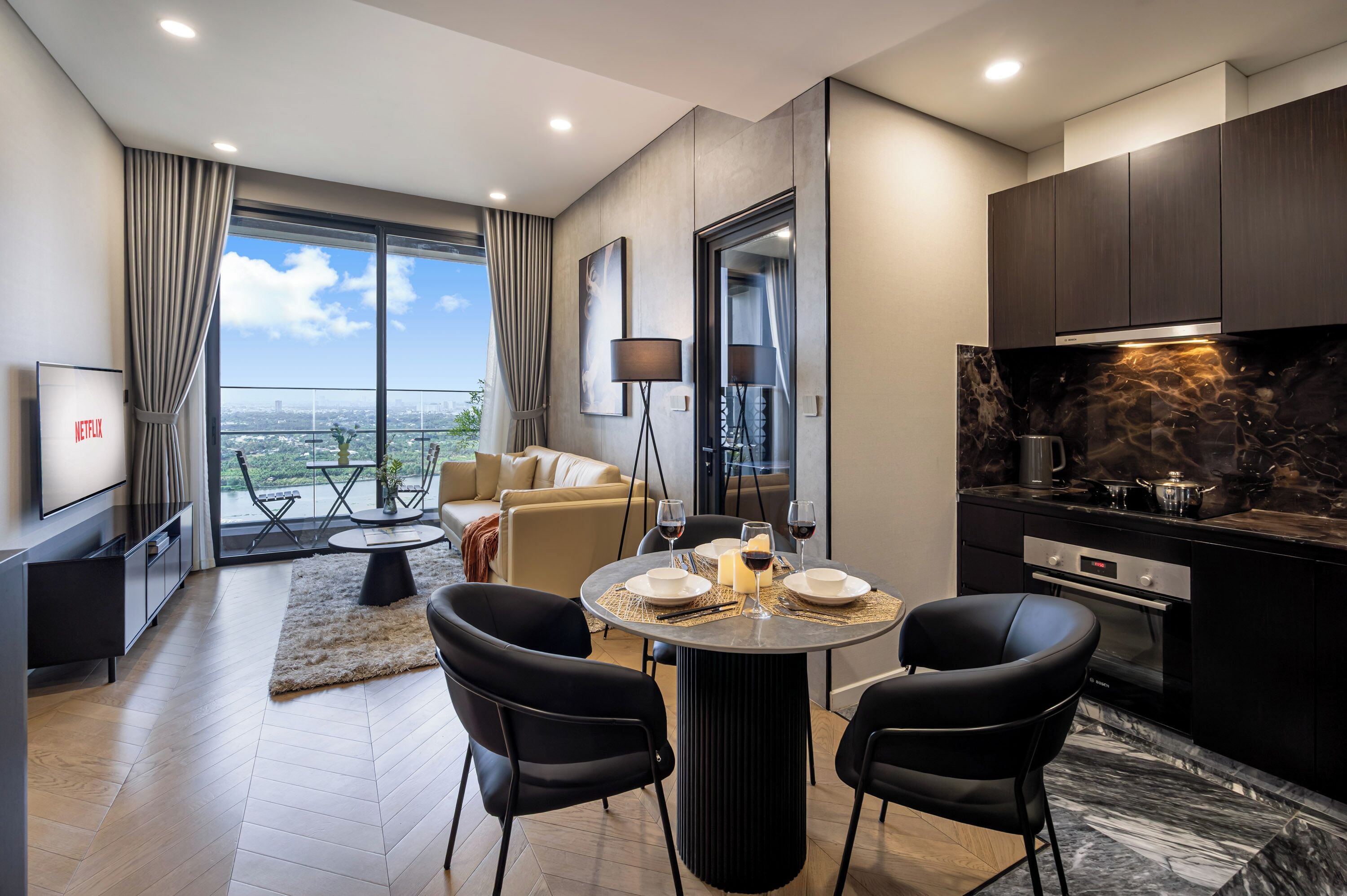 A modern living room with a large window, a dining area with a round table, and a kitchen with dark cabinetry and a marble countertop.