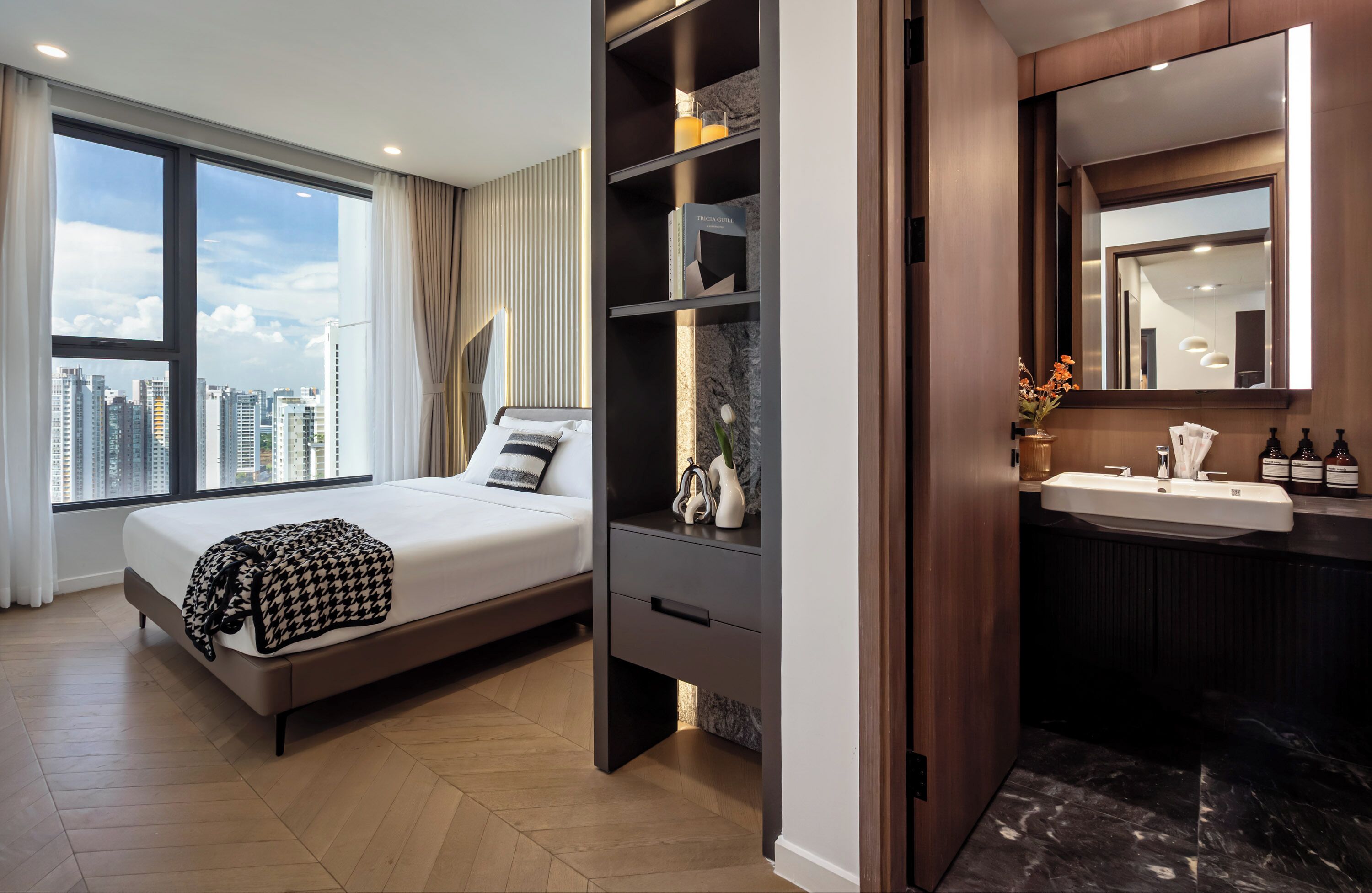 A modern hotel room with a large bed, a built-in wardrobe, a vanity with a mirror, and a view of the city skyline.