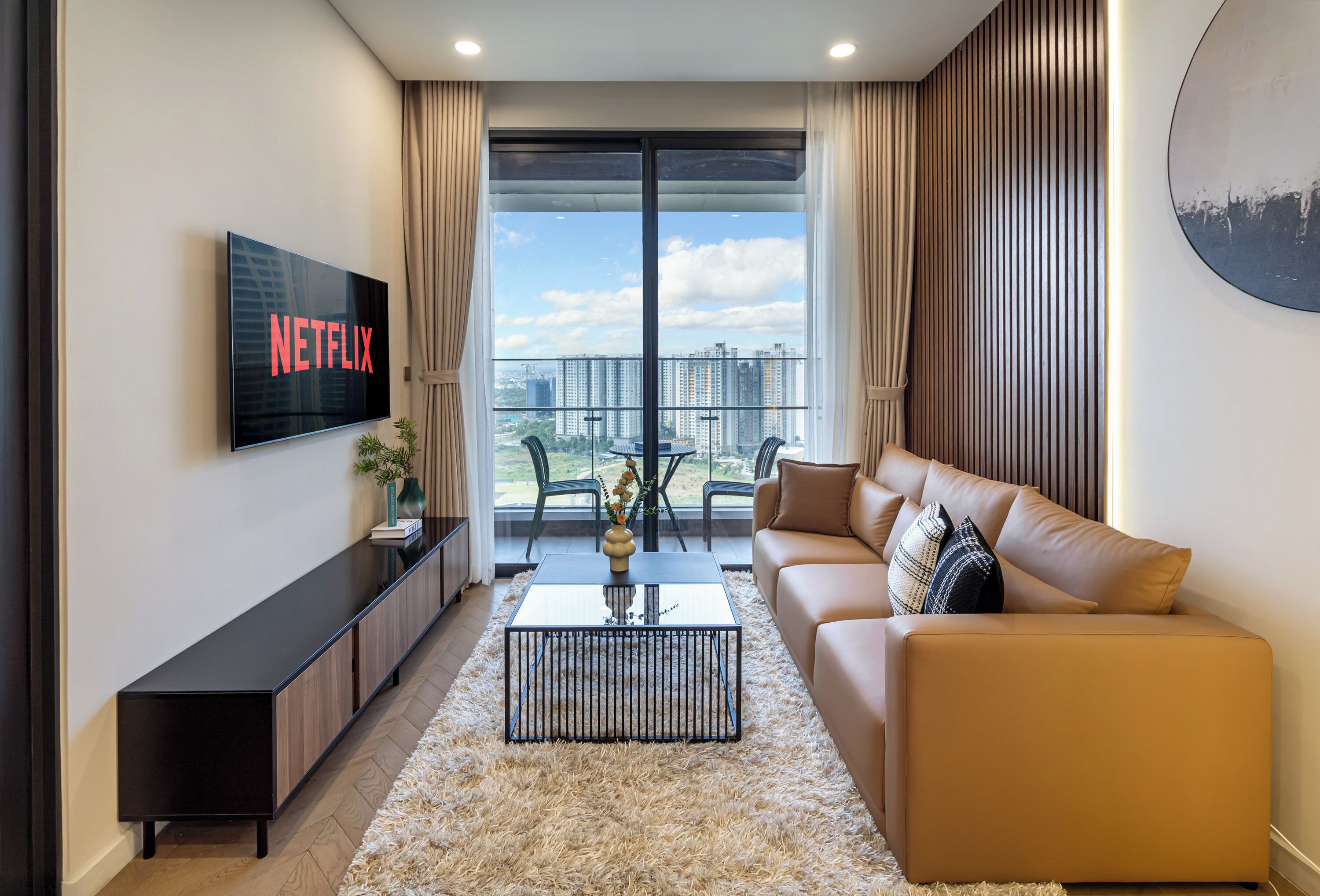 Una sala de estar moderna con un televisor de pantalla plana grande, un sofá, una mesa de centro y vistas a edificios altos a través de ventanas amplias.