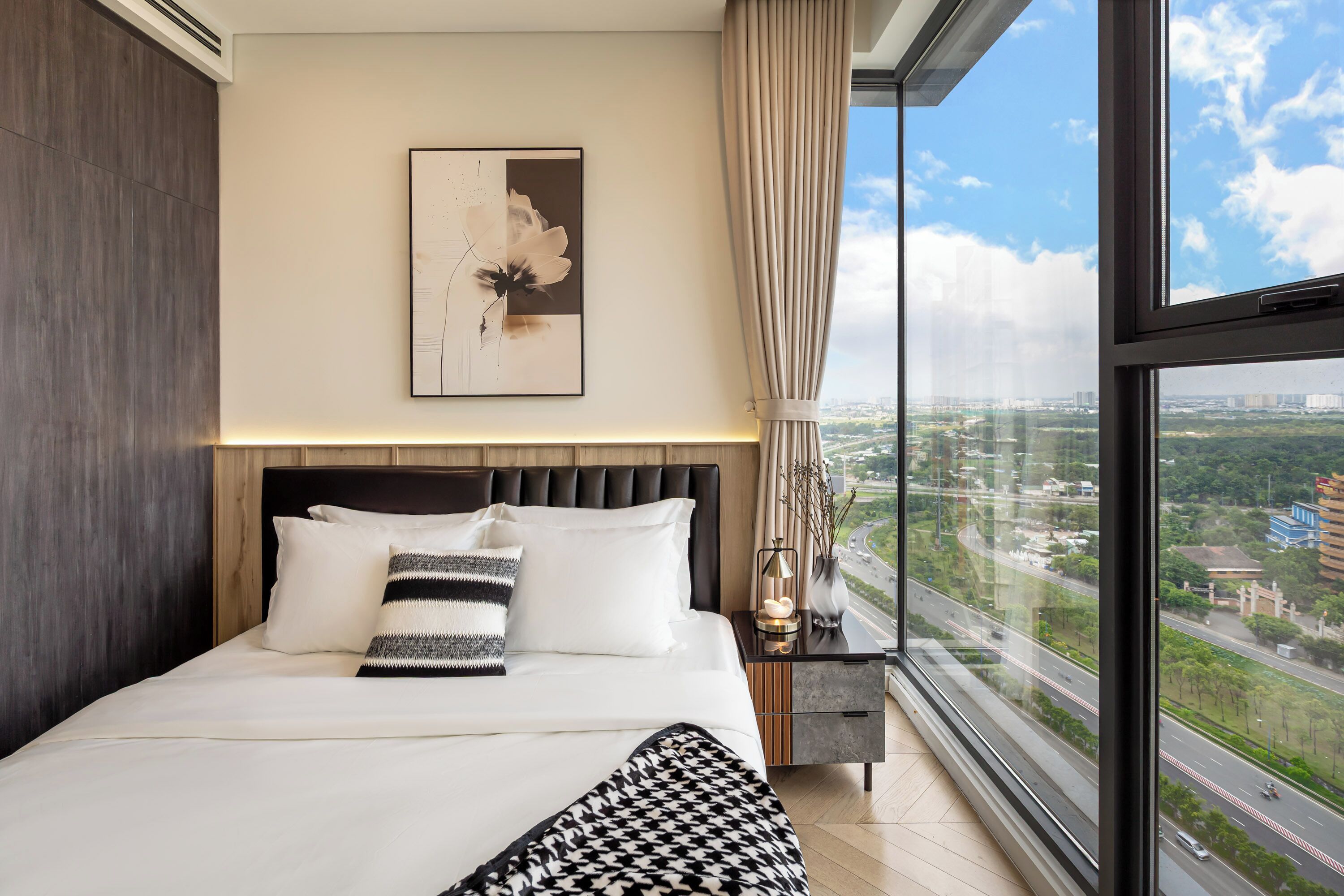 A modern hotel room with a large bed, a city view, a framed artwork on the wall, and a nightstand with a lamp.