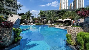 Piscina cercada por palmeiras e espreguiçadeiras, com edifícios altos ao fundo.