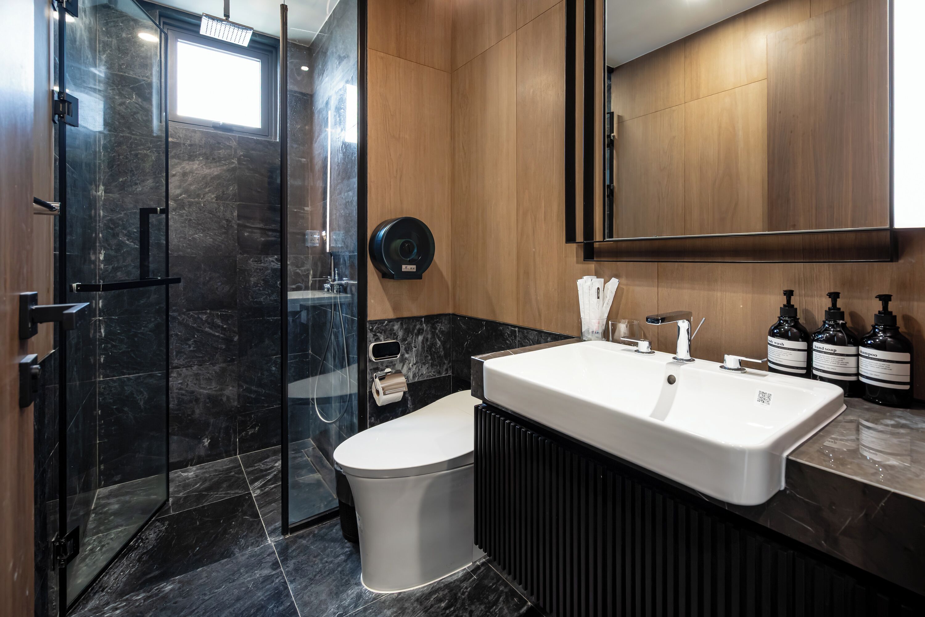Un baño moderno con ducha de vidrio, inodoro blanco y un lavamanos grande con tres botellas de jabón.