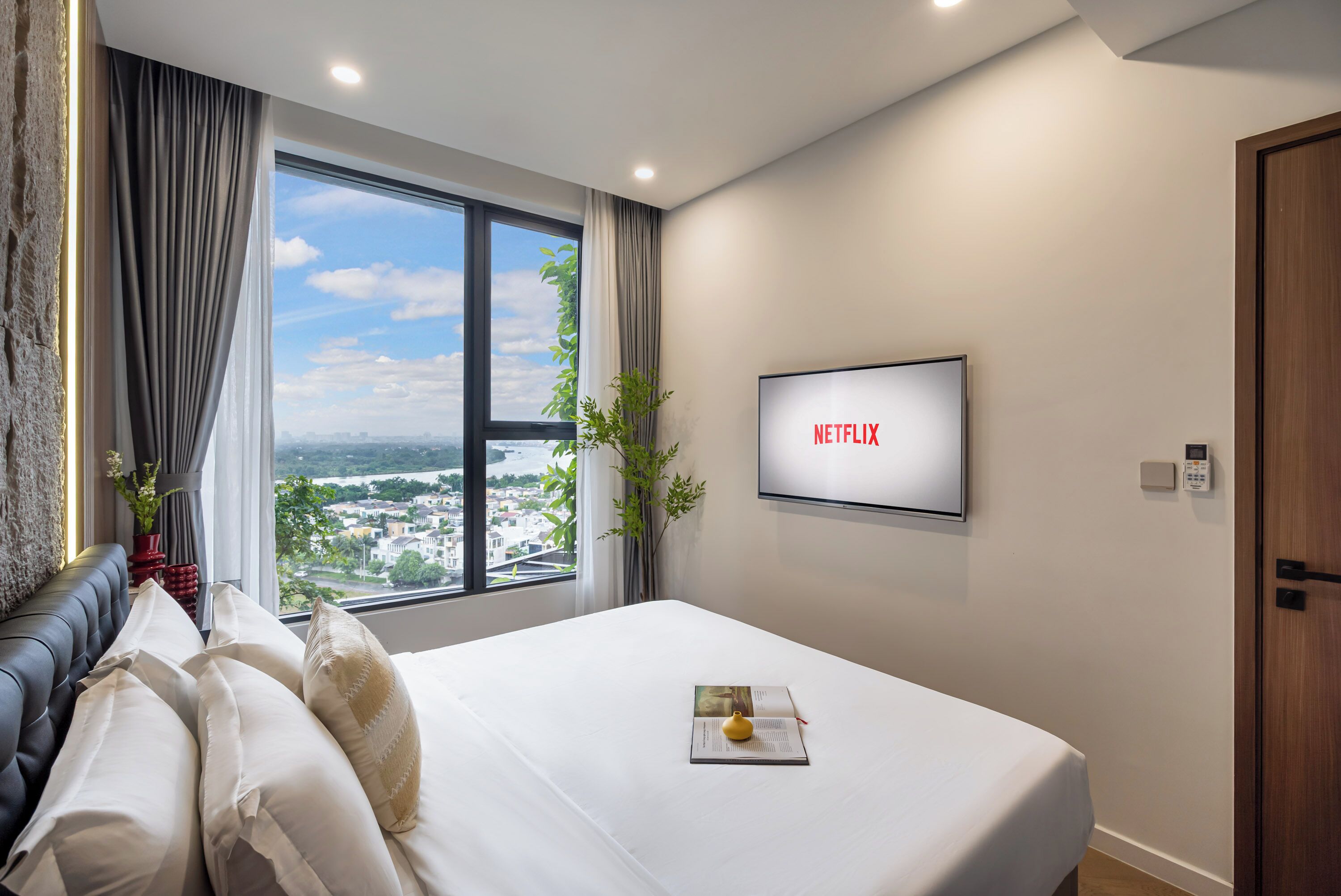 A modern hotel room with a large bed, a view of a cityscape through a window, a wall-mounted TV, and a bedside table with a book and a lamp.