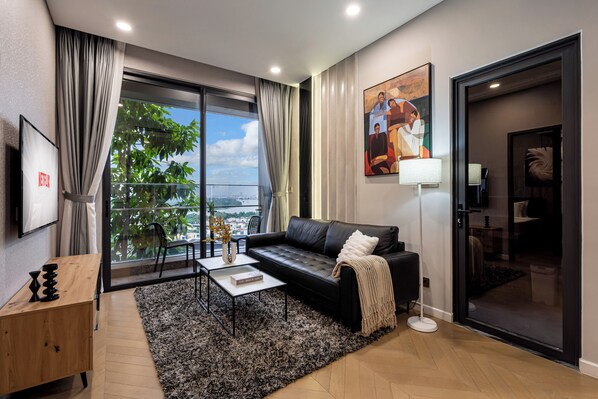 A modern living room with a black leather sofa, a coffee table, and a large window offering a view of greenery and a cityscape. - Lumiere Riverside by TrueStay (Ho Chi Minh City)