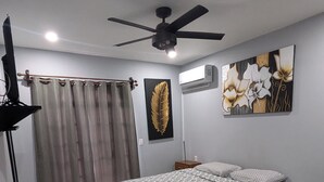 A bedroom with a bed, a ceiling fan, a wall-mounted air conditioner, a curtain, and a painting.