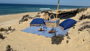 A beach setting with a beach umbrella, a wicker chair, and a blue towel laid out on the sand. - Quinta Amala (Grandola)