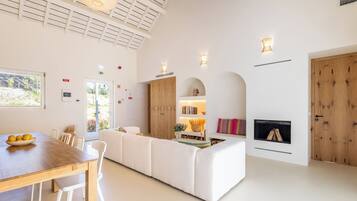 A spacious living area with a wooden dining table, white sofas, a fireplace, and a large window.