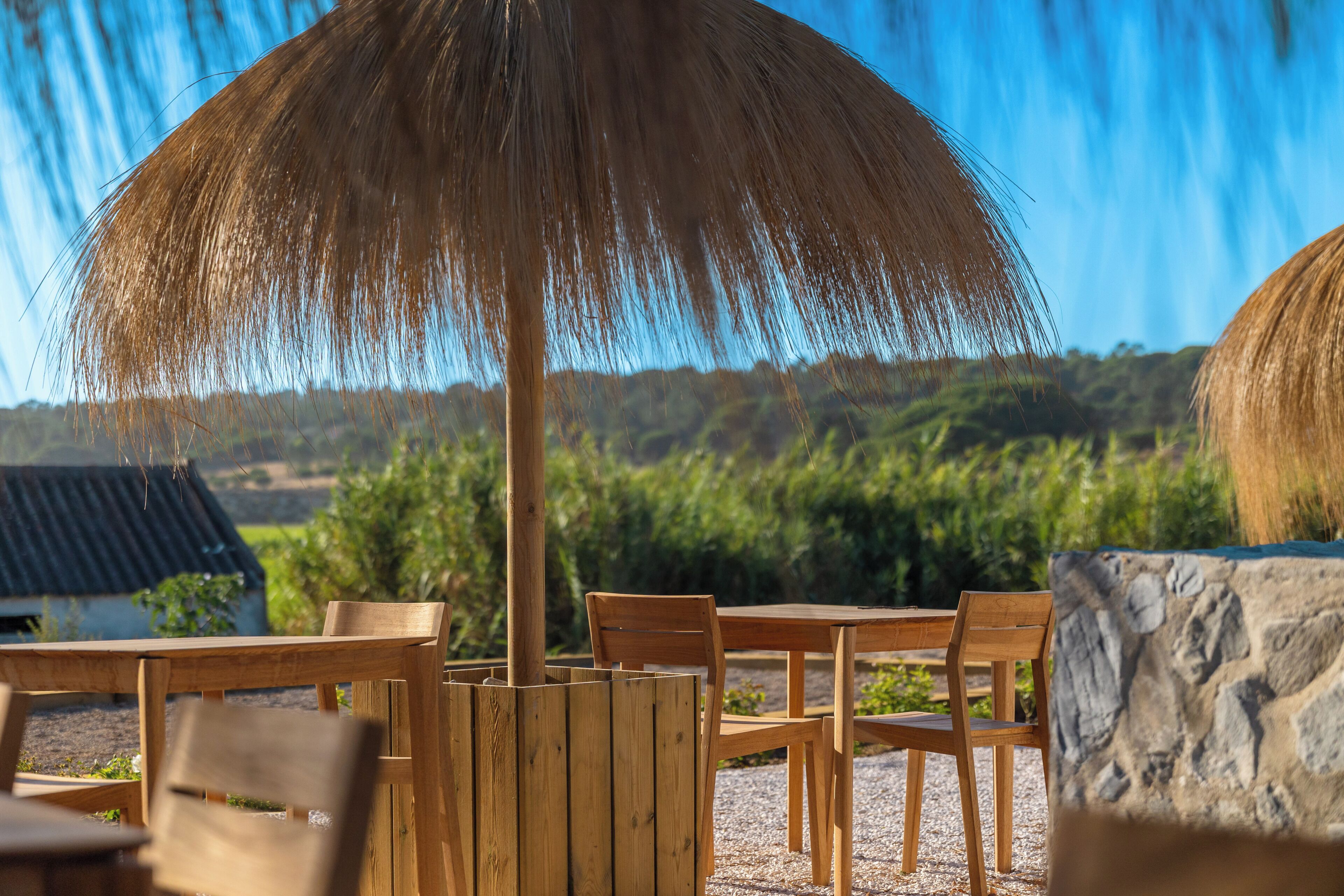 A wooden outdoor seating area with a thatched umbrella, stone wall, and a view of green hills.