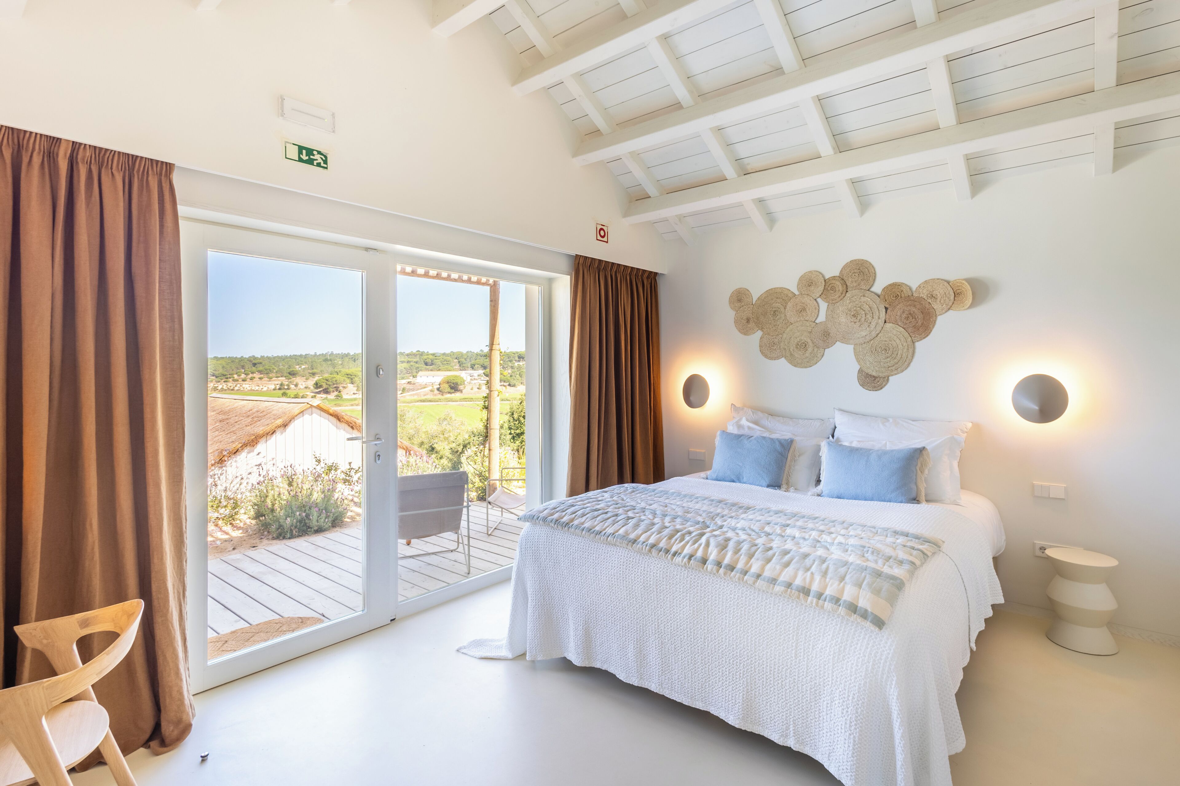 A bedroom with a bed, a chair, a bedside table, a wall-mounted light, and a view through a sliding door.