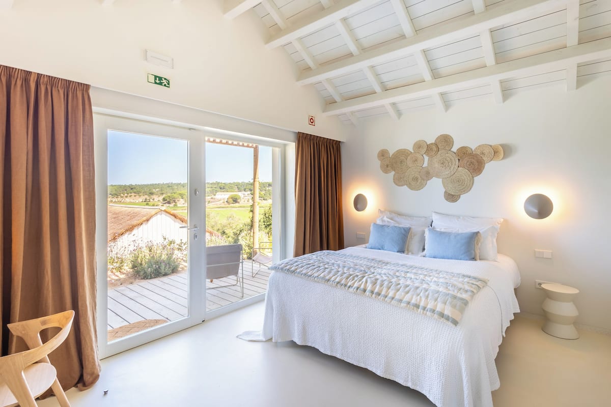 A bedroom with a bed, a chair, a bedside table, a wall-mounted light, and a view through a sliding door.
