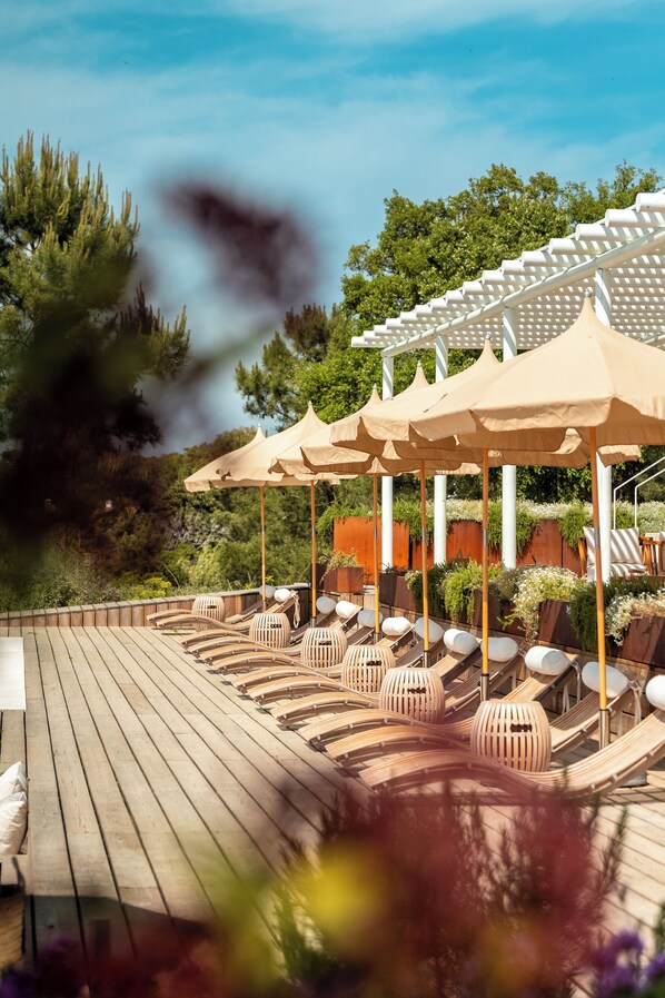 A wooden deck with lounge chairs and beige umbrellas, surrounded by greenery and trees.