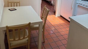 Eine kleine Küche mit weißen Schränken, einem weißen Kühlschrank und einem Gasherd. Ein Esstisch mit Stühlen befindet sich im Vordergrund.