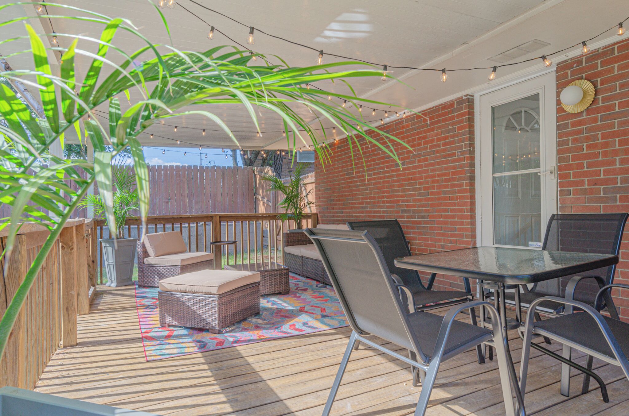 A covered patio with a brick wall, a glass table, metal chairs, wicker furniture, and a colorful rug.