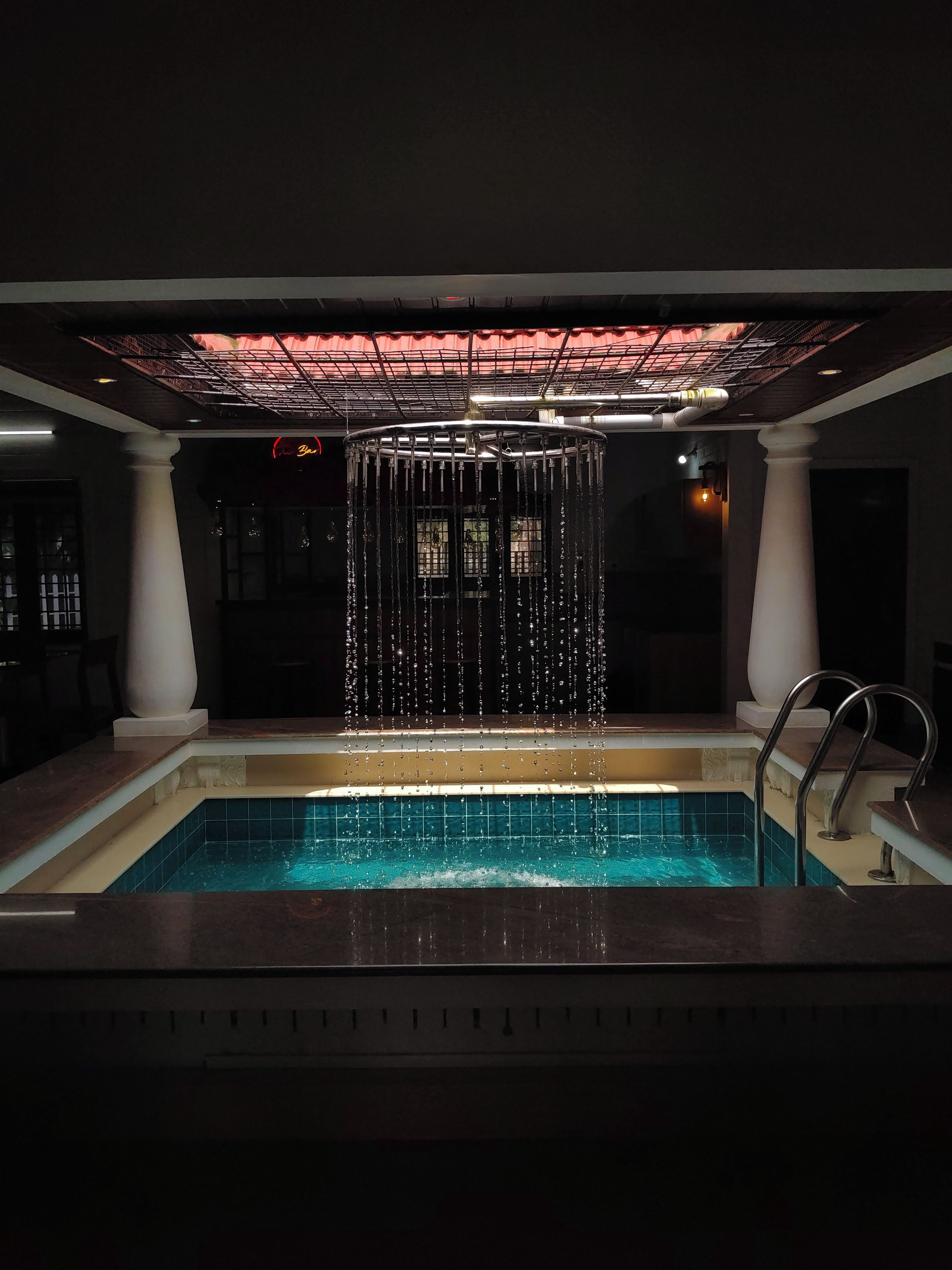 Une piscine agrémentée d’un élément décoratif aquatique au centre, entourée de colonnes et dotée d’un sol carrelé.