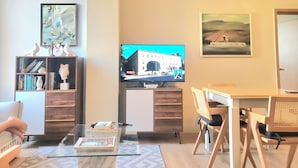 A living room with a flat-screen TV, a bookshelf, a glass coffee table, a desk with a chair, and a painting on the wall.