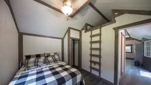 A bedroom with a checkered bedspread, wooden beams, a ladder, and a view into another room. - Tiny House w/Creek and Eagles (Millersburg)