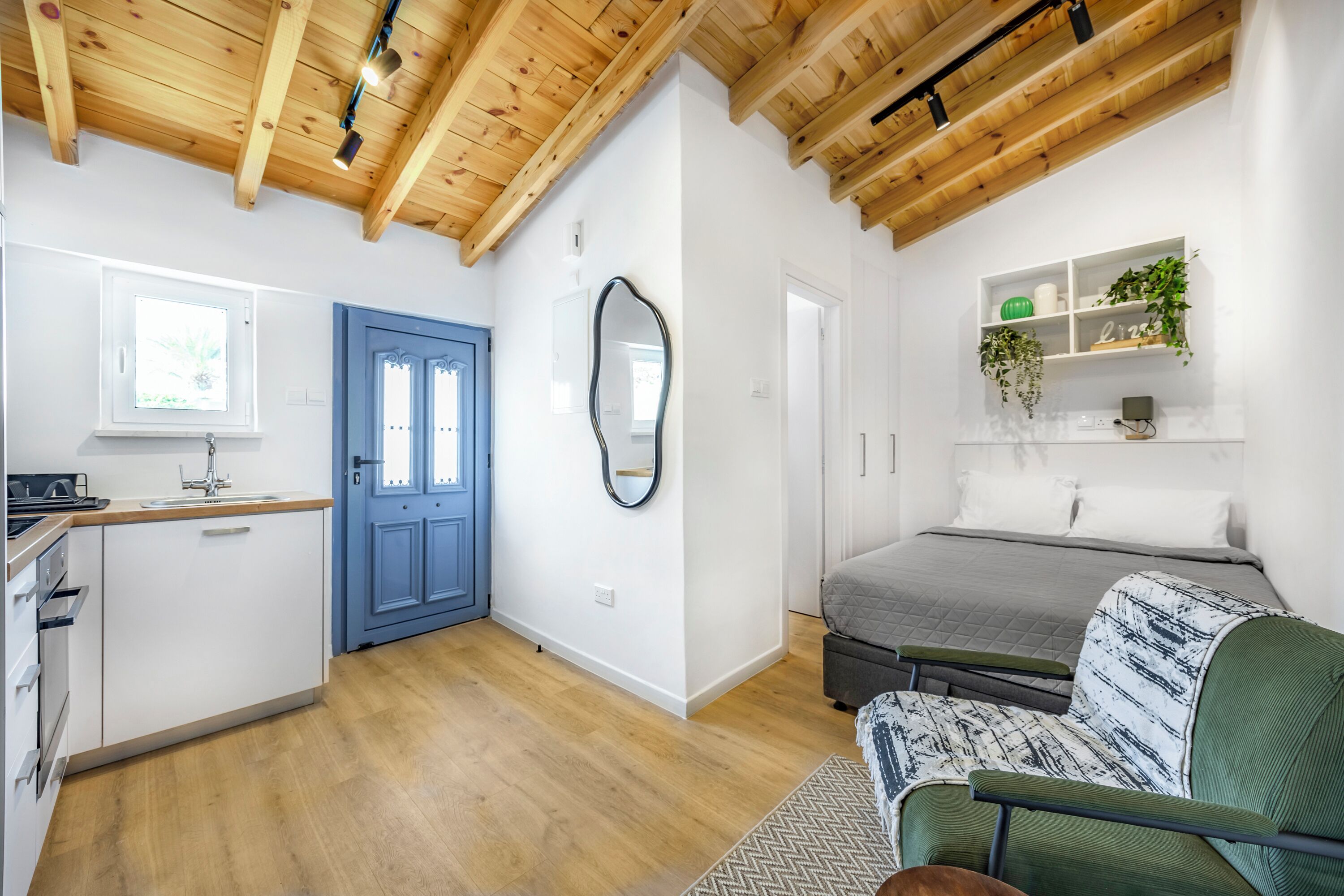 A compact living space with a kitchenette, a blue door, a mirror, a bed with a patterned blanket, and a shelf with plants.