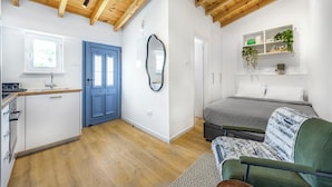 A compact living space with a kitchenette, a blue door, a mirror, a bed with a patterned blanket, and a shelf with plants. - Olga Apt 4- Lovely Studio on Ground Floor (Limassol)