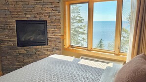 Un dormitorio con pared de piedra, una chimenea, un ventanal con vistas al mar y una cama con ropa blanca.