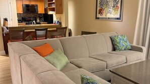 Una sala moderna con un amplio sofá modular en forma de L, una mesa de centro y un comedor con armarios de madera y una cocina al fondo.