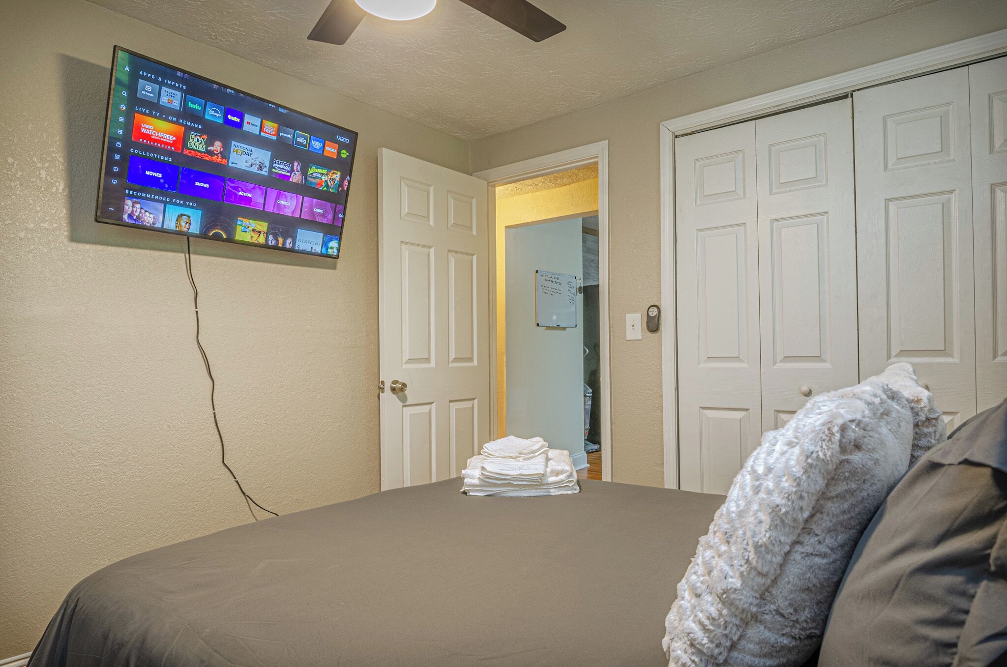 A bedroom with a bed, a television mounted on the wall displaying a streaming service interface, and a door leading to another room.