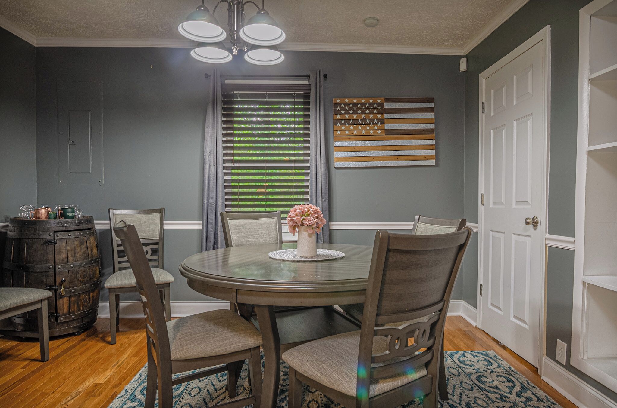 A dining area with a round table, four chairs, a vase with flowers, a wooden barrel, and a wall with an American flag.