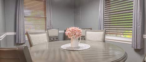 Una mesa redonda de comedor con cuatro sillas, un jarrón de flores rosadas y un mantel individual con textura.