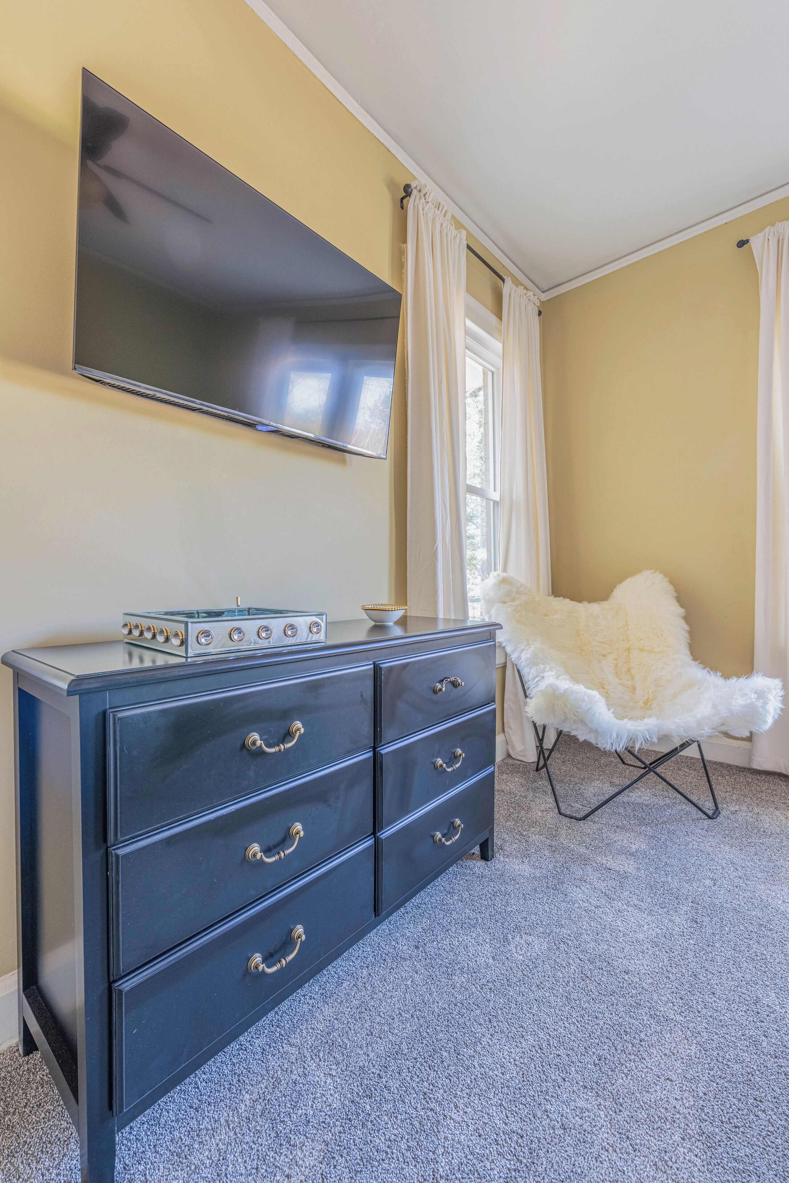 A room with a flat-screen TV mounted on the wall, a dark blue dresser with drawers, a fluffy white chair, and a window with curtains.