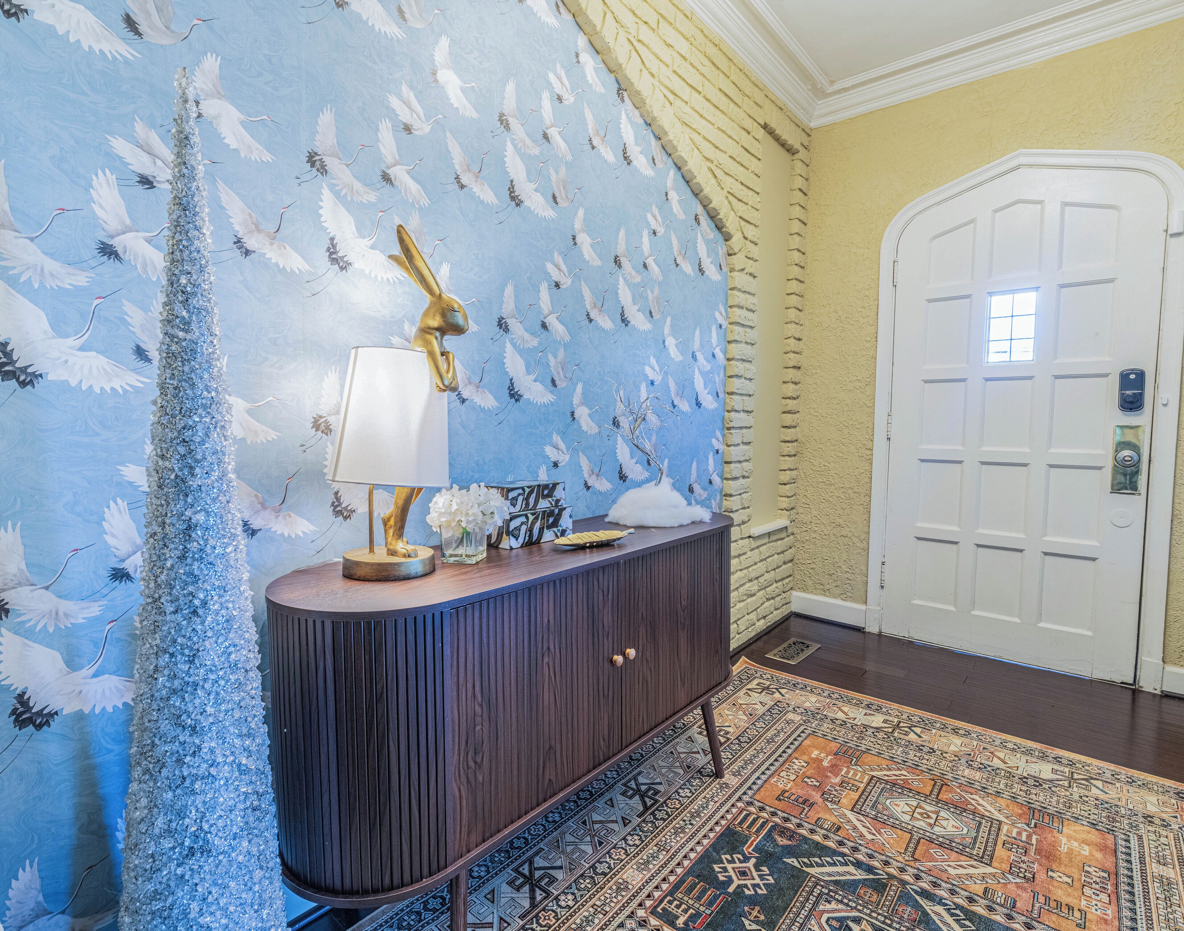 A hallway with a decorative wall featuring white birds and a golden statue, a wooden console table with a lamp, and a patterned rug.
