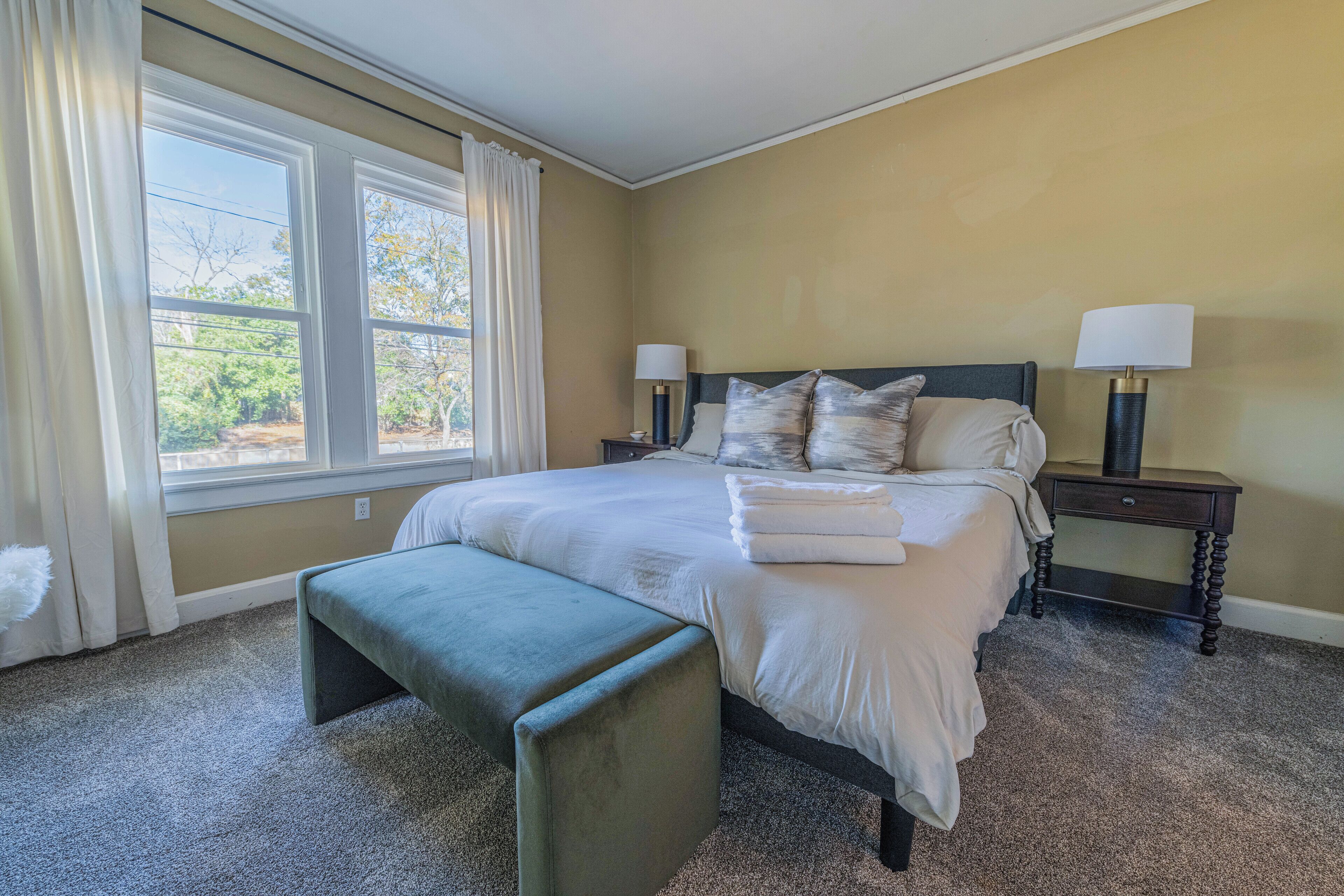 A bedroom with a large bed, two bedside tables with lamps, a bench, and a window with curtains.