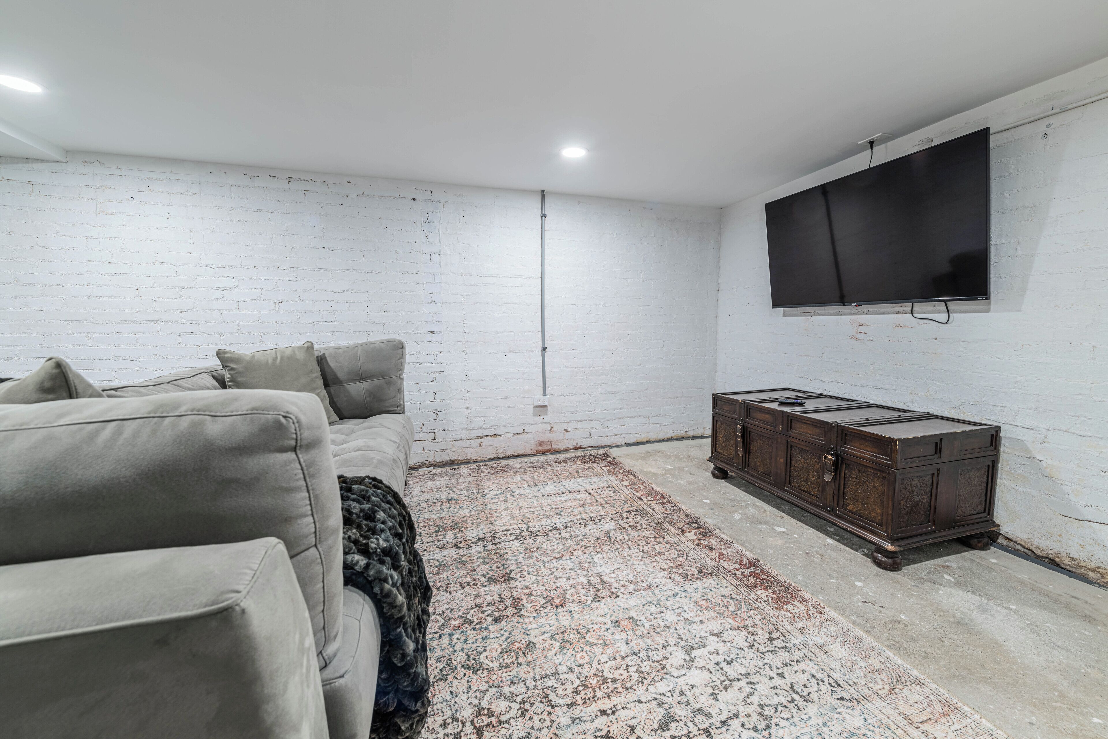 A room with a white brick wall, a large flat-screen TV mounted on the wall, a grey sofa, a patterned rug, and a wooden cabinet.