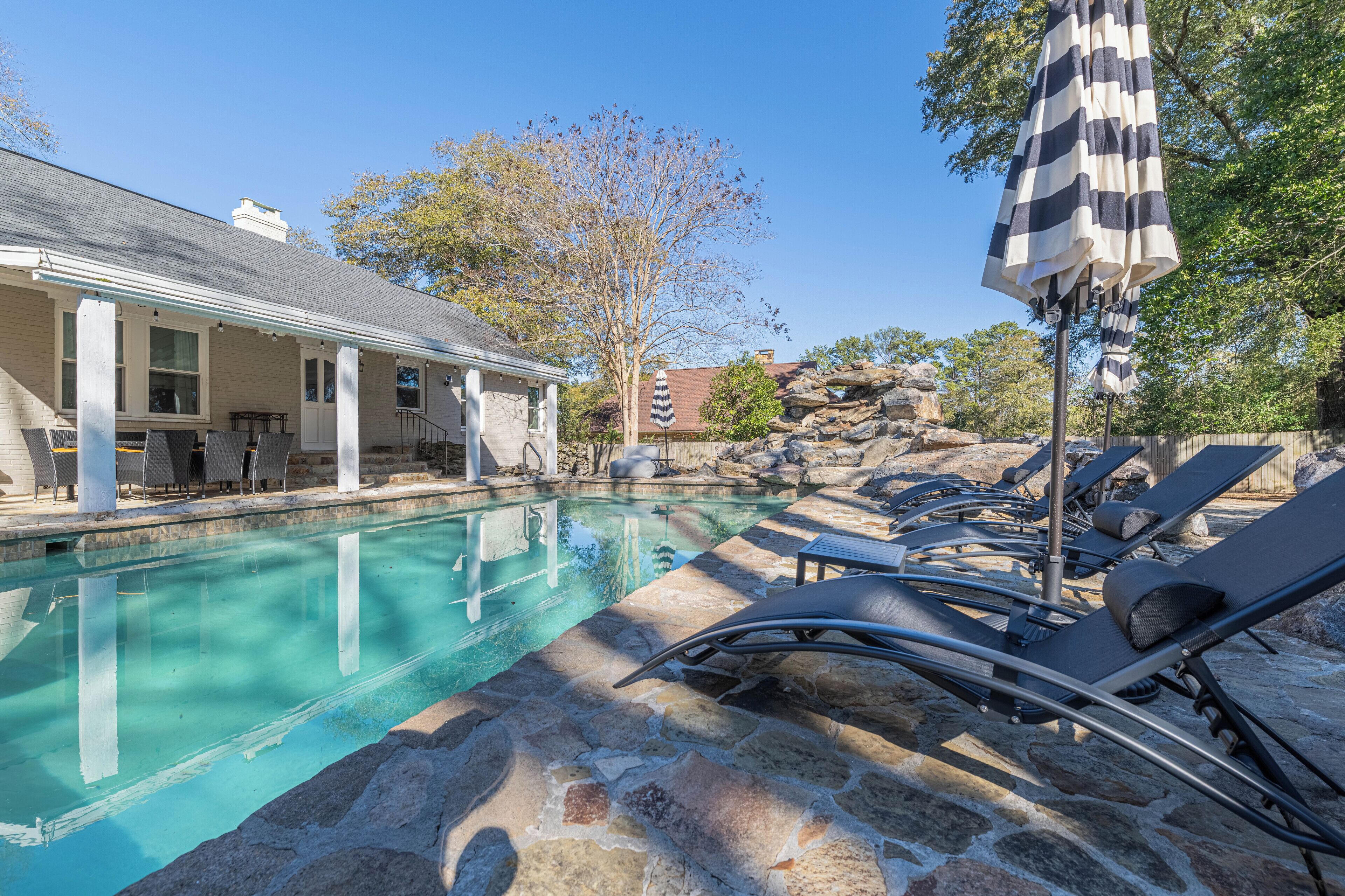 A swimming pool with lounge chairs and an umbrella, surrounded by stone tiles and a building with a white facade.