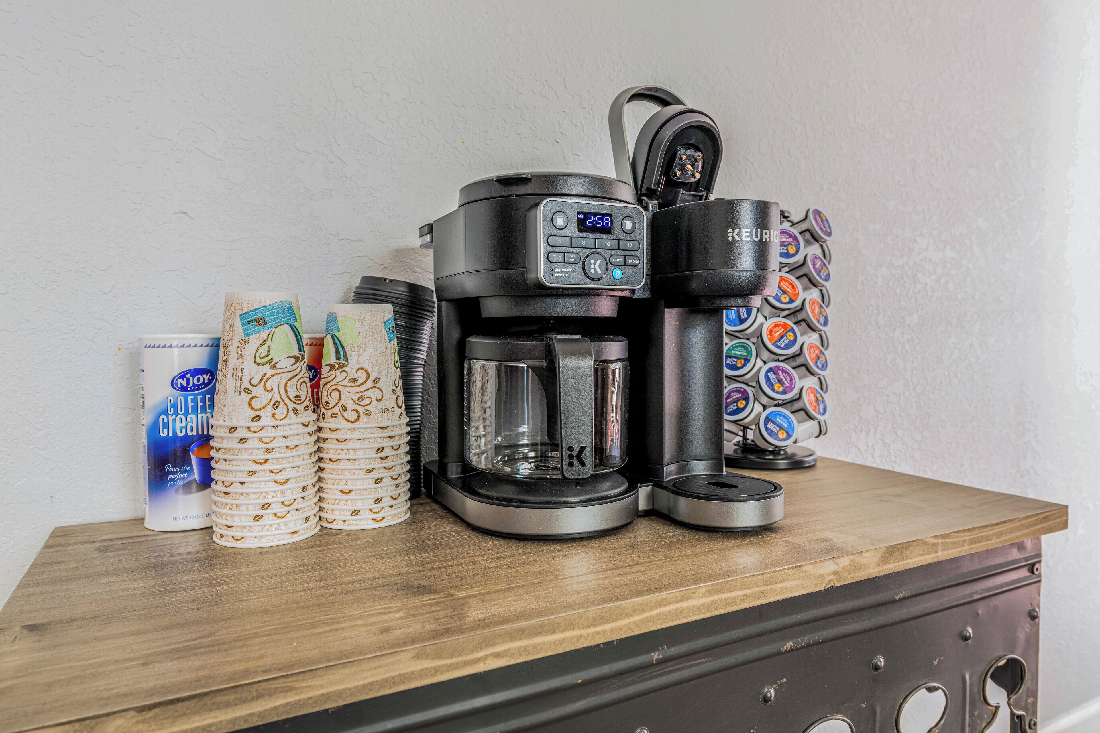 A coffee maker, a stack of disposable coffee cups, and a coffee creamer can on a wooden surface.