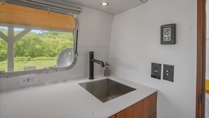 A modern kitchen with a stainless steel sink, a window with a view of greenery, and a thermostat on the wall.