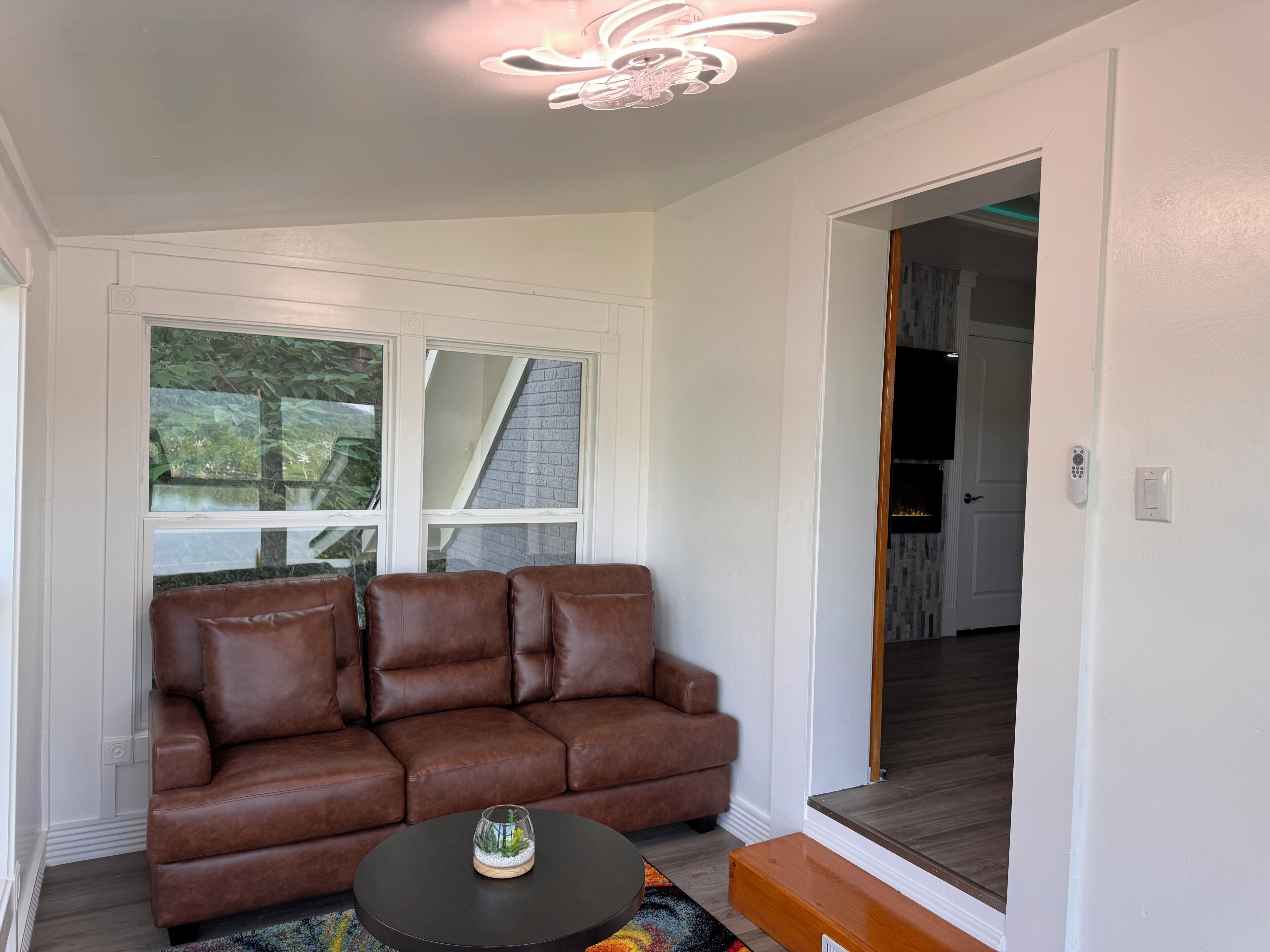 A living room with a brown leather sofa, a round coffee table, and a glass-top table with a decorative item. There are two windows with a view of greenery outside, and a door leading to another room.