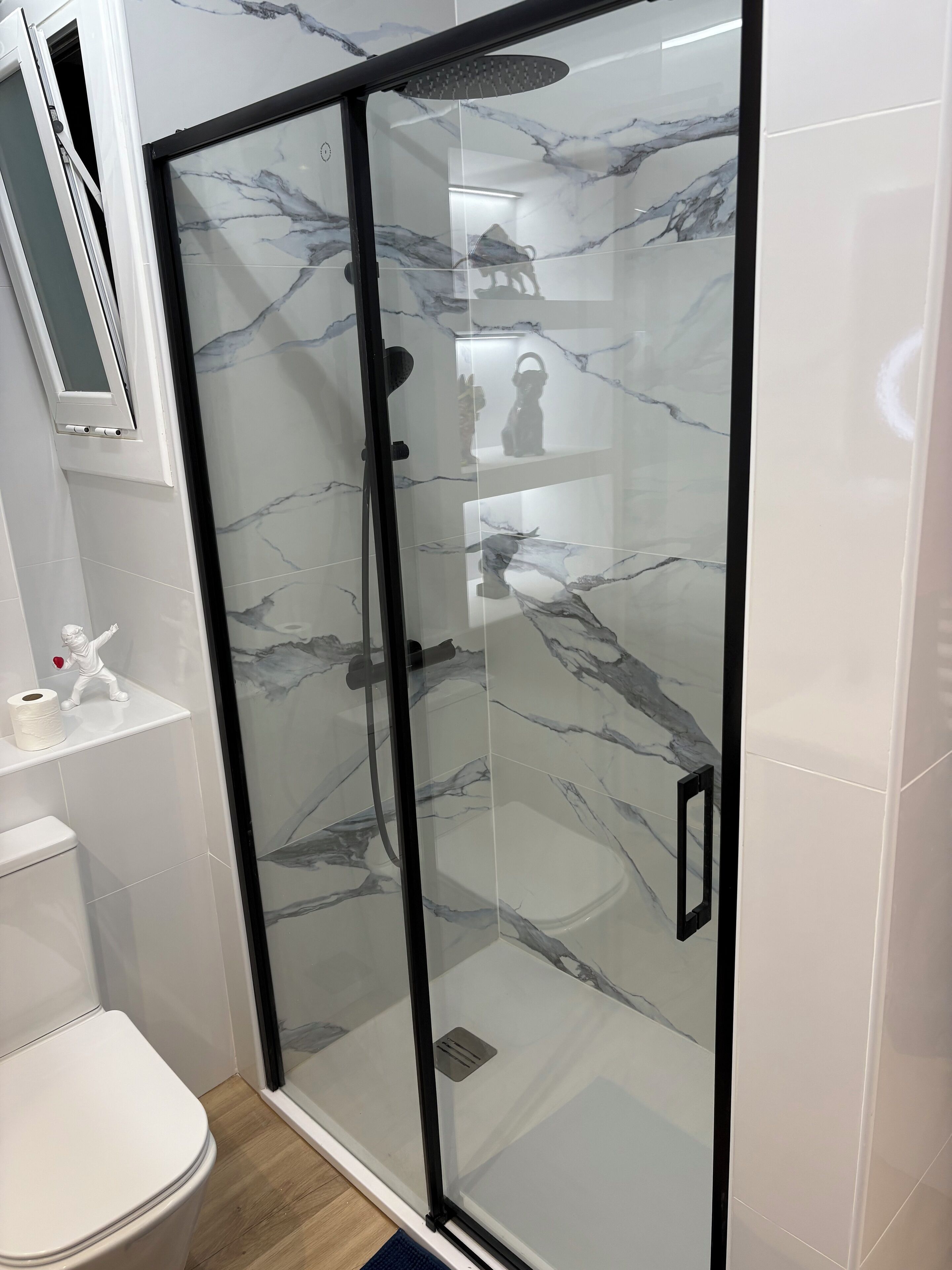 Ein modernes Badezimmer mit einer Duschkabine, Marmorfliesen, einer Toilette und einer kleinen Ablage für Pflegeprodukte.